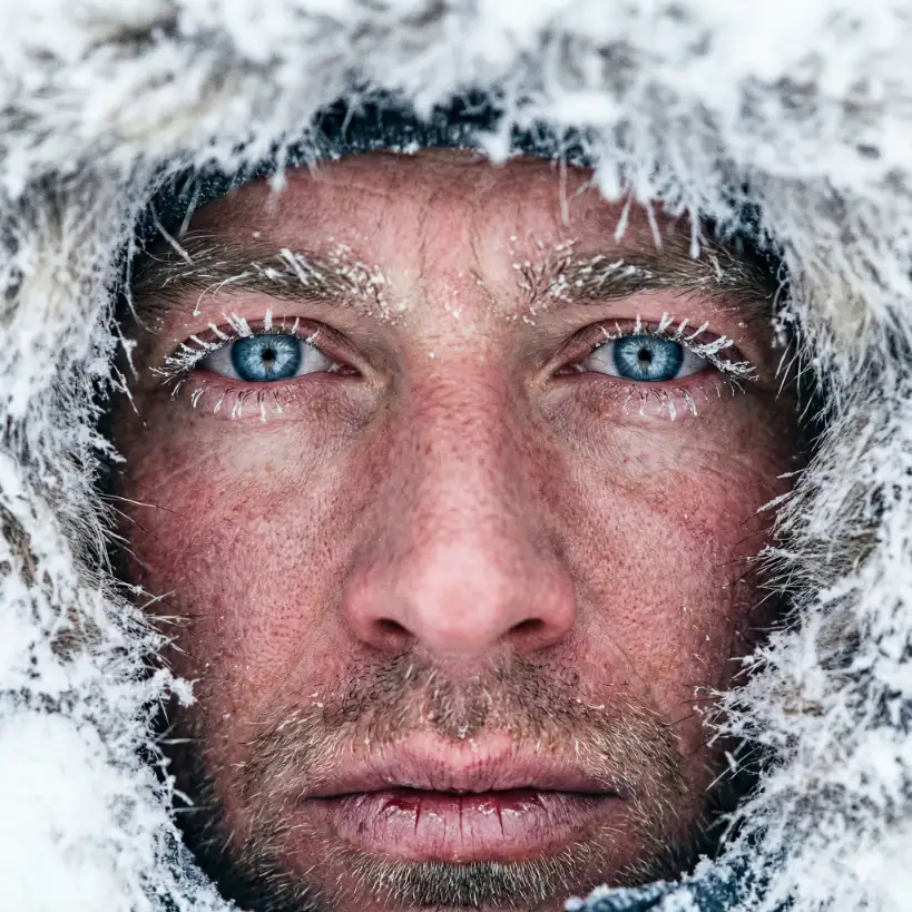 Create an extreme close-up, hyper-realistic portrait of a mountaineer in a snowy environment. The subject is wrapped in a thick parka with a fur hood. The focus is entirely on the eyes and nose. There are tiny ice crystals frozen onto their eyelashes and eyebrows. The skin is windburned and ruddy from the cold. The lighting is the incredibly bright, diffuse white light typical of a snowy day, reflecting in their irises. No background is visible other than the blur of white snow and fur. Capture the texture of chapped lips. Aspect ratio: 1:1.