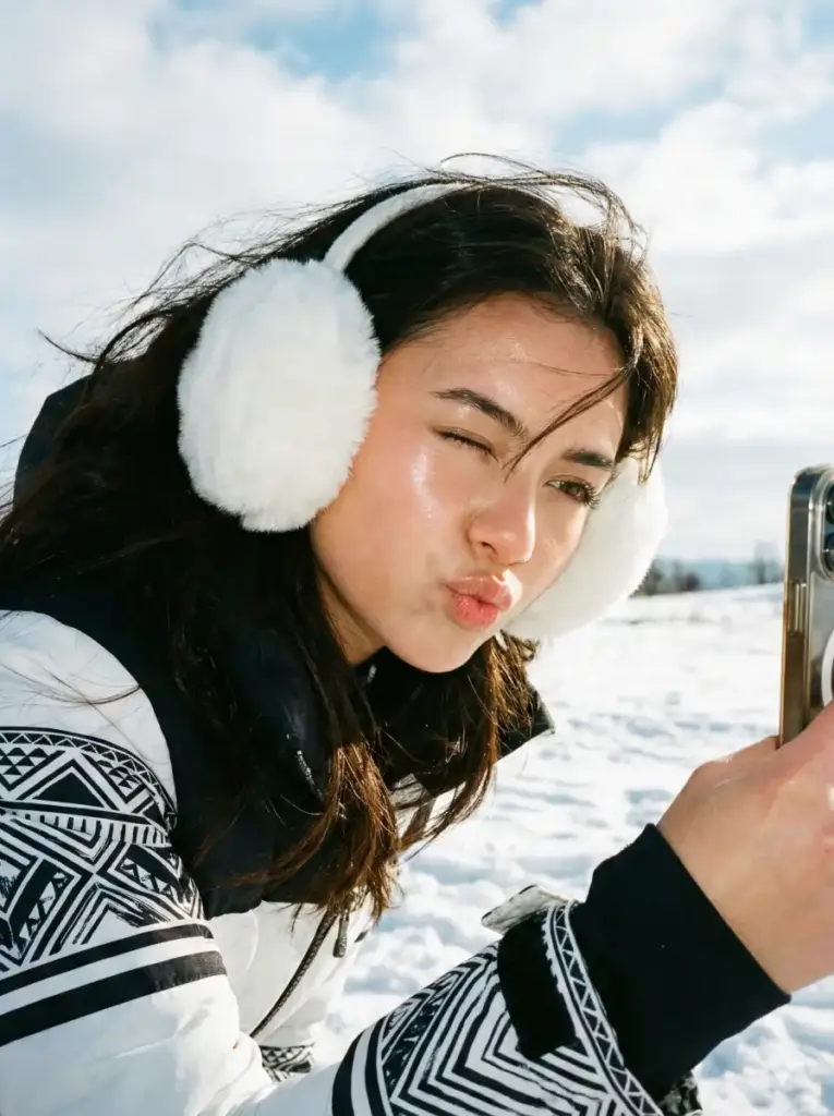 Create photo of a young woman takes a playful winter selfie outdoors, leaning slightly toward the camera as she puckers her lips in a fun, affectionate expression. She wears large, fluffy white earmuffs that frame her face and contrast with her long, flowing dark hair, which is blown gently by the cold breeze. Her skin glows naturally in the crisp daylight, with sunlight illuminating her features and casting soft highlights along her hair. She's dressed in a winter jacket decorated with black-and-white geometric patterns on the sleeves, adding a stylish and cozy touch. Behind her, the sky is bright and clear with soft wisps of clouds, and hints of snowy landscape can be seen near the horizon. The overall vibe is cheerful, lively, and candid. Use aspect ratio 3:4.