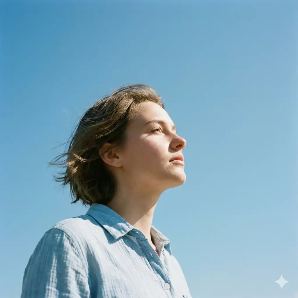 Create a cinematic, minimalist portrait photographed from a low angle of a young woman standing outdoors beneath a vast, cloudless sky. The subject is shown in side profile, gazing upward toward the sun with a calm, contemplative expression. Soft natural daylight illuminates their face, creating gentle highlights along the cheek and nose while leaving subtle shadows under the jawline. Their short, slightly tousled hair moves lightly in the breeze. They are wearing a loose, light-blue button-up shirt with relaxed folds and natural fabric texture. The composition emphasizes negative space, with the expansive blue sky occupying most of the frame, evoking a sense of openness, freedom, and quiet introspection. Use aspect ratio 1:1.