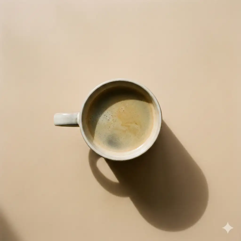 Create a minimalist top-down still life of a single coffee mug placed against a smooth, warm beige background. The mug is centered slightly above the middle of the frame, viewed directly from overhead. Inside the cup, freshly poured coffee shows subtle crema or foam texture on the surface, with gentle tonal variation. Soft, directional natural light enters from one side, casting a long, smooth shadow of the mug diagonally across the background. The shadow is clean-edged yet soft, adding depth and visual balance without clutter. The handle of the mug is visible, contributing to a simple, graphic silhouette. Use aspect ratio 1:1.