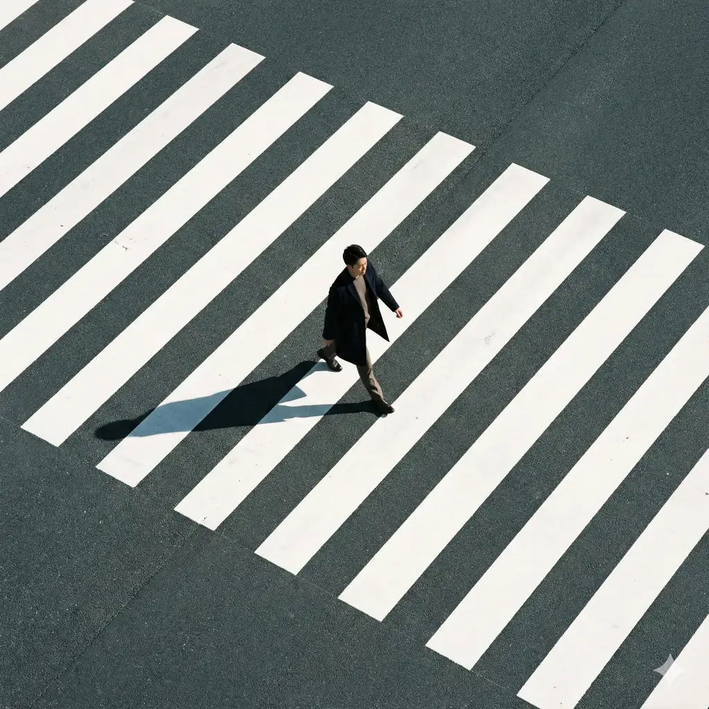 Create a high-angle, top-down urban minimalist photograph of a lone pedestrian crossing a wide city crosswalk. The scene is dominated by bold, parallel white zebra stripes cutting diagonally across dark gray asphalt, creating a striking graphic pattern. The road surface is textured and matte, with subtle wear and tonal variation. At the center of the composition, a single figure walks confidently across the stripes, captured mid-step. The person wears a dark, tailored coat and minimal, neutral clothing, contrasting sharply against the bright white lines. Their long shadow stretches diagonally across the crosswalk, adding depth, motion, and a sense of late-morning or afternoon sunlight. Use aspect ratio 1:1.