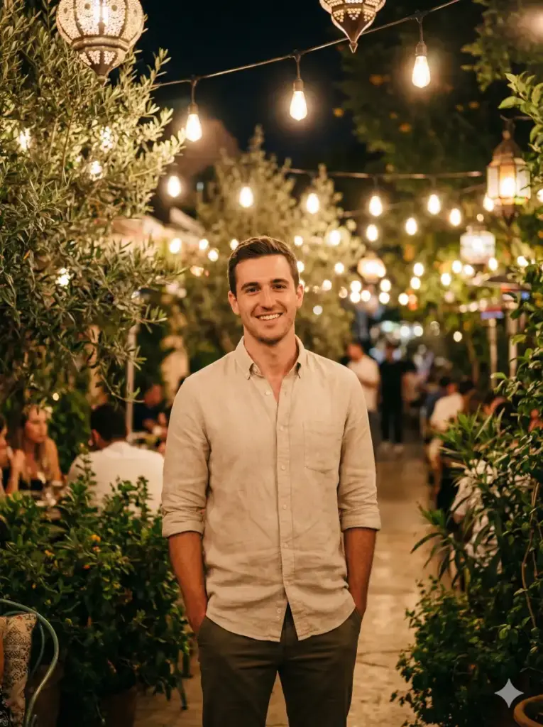 Create an outdoor night-time lifestyle portrait of a young adult man standing in a cozy café or garden setting illuminated by warm hanging lights. He stands in a relaxed, natural posture with a gentle, confident smile directed toward the camera. His outfit is casual-smart, featuring a light-colored button-up shirt with sleeves rolled slightly for an easy, approachable look. Soft ambient lighting from decorative lamps and string lights surrounds him, creating warm bokeh and a festive atmosphere. Green plants and foliage frame the scene, adding depth and visual balance. The lighting is warm and flattering, with even exposure on the face and soft shadows. The background remains lively yet softly blurred to maintain focus on the subject. The image features shallow depth of field, realistic skin tones, subtle grain, and a warm editorial color grade. Photorealistic lifestyle photography, natural expressions, evening ambiance, cozy mood, no harsh lighting, aspect ratio 3:4.