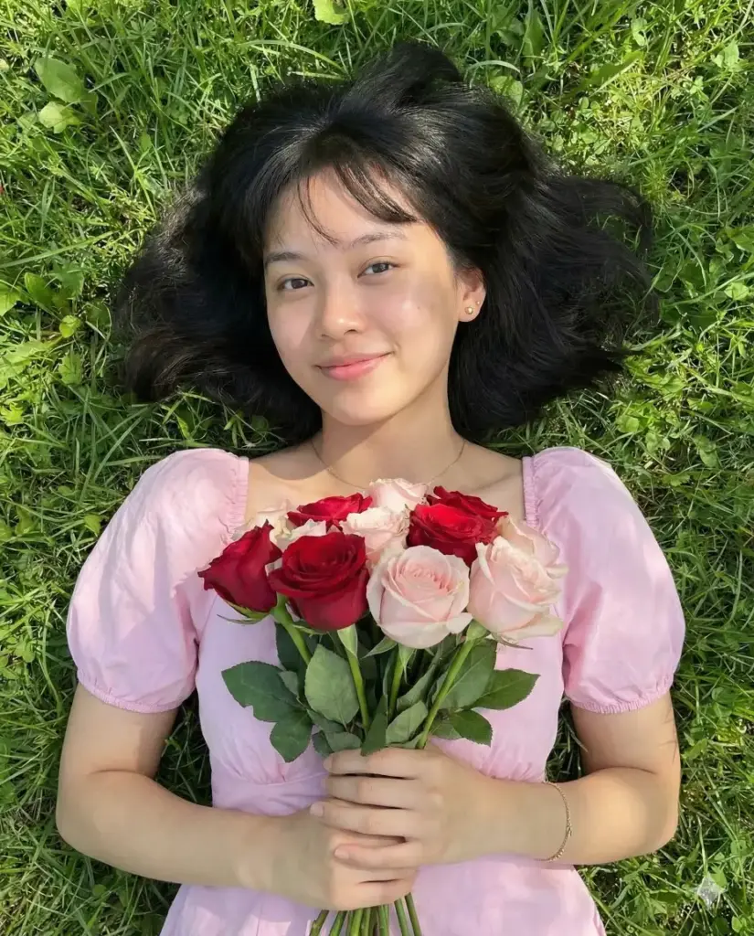 Create a photorealistic romantic lifestyle portrait of a young woman lying on her back on lush green grass, captured from a top-down perspective and framed from the chest up. She gently holds a bouquet of fresh roses against her chest, featuring a mix of deep red and soft blush-pink blooms with visible stems and leaves. She wears a soft pastel pink dress with short puff sleeves, giving the scene a delicate, feminine feel. Her dark hair is loose and spread naturally on the grass, with light bangs framing her face. She gazes directly at the camera with a calm, tender expression and a subtle smile, conveying warmth and quiet intimacy. Her makeup is minimal and natural with realistic skin texture and a soft glow, and she wears delicate jewelry including small earrings, a fine necklace, and a simple bracelet. Lighting is bright natural daylight with even illumination and gentle shadows, preserving true-to-life colors and fine detail in the grass and flowers. The background is fully filled with rich green grass, clean and natural with no empty areas or artifacts. The overall mood is soft, romantic, and serene, like a candid spring afternoon moment. High resolution, realistic lens depth, no distortion, no text. Aspect ratio 4:5.
