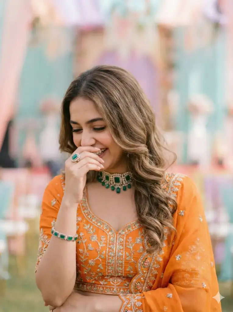 Create a close-up festive lifestyle portrait of a young adult woman captured in a candid, joyful moment. She tilts her head slightly downward while smiling softly, with one hand raised to partially cover her face, conveying a shy, genuine laugh. She wears a vibrant orange traditional outfit with delicate embroidery, styled elegantly for a celebration. Her jewelry includes a green gemstone choker necklace, a matching bracelet, and a subtle ring that adds refined detail. Her hair is styled neatly with soft waves cascading over one shoulder. Bright natural daylight illuminates her face evenly, creating smooth skin tones and gentle highlights. The background remains softly blurred with light pastel hues to maintain focus on emotion and expression. The image features shallow depth of field, subtle grain, balanced color, and a soft editorial finish. Photorealistic festive portrait photography, natural light, candid emotion, elegant styling, no harsh shadows, aspect ratio 3:4.
