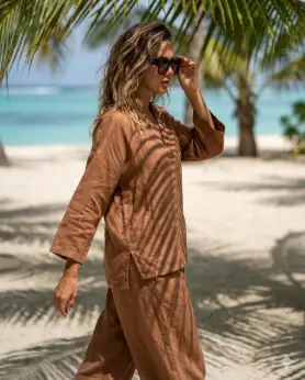 Create a stylish beach lifestyle photo of a woman walking slowly along the sand under palm tree shadows, natural upright posture with one arm swinging and the other adjusting sunglasses, wearing a coordinated beach outfit with flowing fabric, soft waves in hair from ocean breeze, relaxed confident facial expression, sunlight filtering through palm leaves creating patterned shadows, camera shot from a side angle at waist height, tropical background softly blurred, clean Instagram travel aesthetic, high realism photography. Use aspect ratio 4:5.
