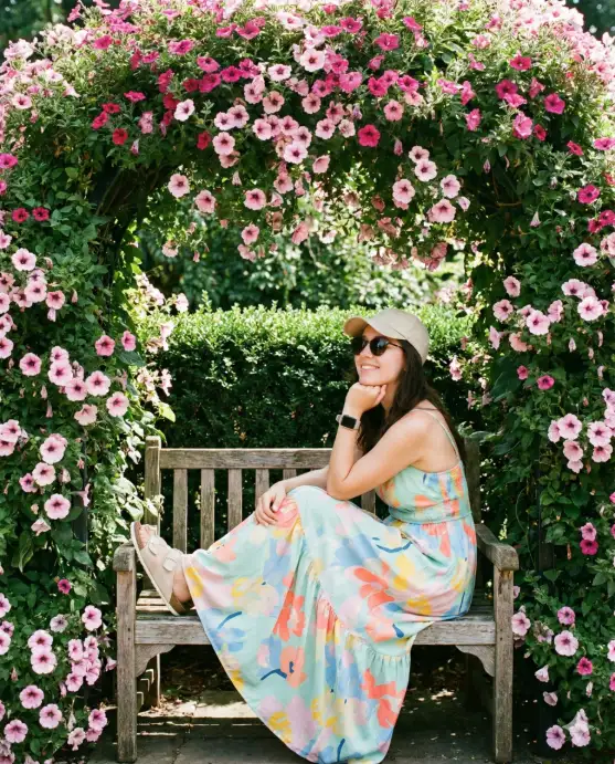 Create a vibrant, whimsical garden portrait of a young woman seated beneath a lush floral arch overflowing with pink and magenta petunias. The archway is densely covered in cascading blossoms, soft pink petals with darker magenta centers, framed by rich green foliage that creates a natural canopy around her. The flowers spill down both sides, forming a romantic, storybook-like entrance. She sits gracefully on a simple bench beneath the arch, one leg slightly lifted in a relaxed, playful pose. Her posture is casual yet elegant, with one elbow resting on her knee and her hand lightly touching her chin as she gazes off to the side with a soft, content smile. She wears a flowing maxi dress in pastel tones,mint, sky blue, coral, yellow, and pink, adorned with large abstract floral patterns. The fabric is lightweight and airy, gently draping around her legs. On her feet are white slip-on shoes with a casual, comfortable style. She accessorizes with a light beige baseball cap, dark sunglasses, and a smartwatch on her wrist, blending modern touches with the romantic garden setting. Behind her, a neatly trimmed green hedge forms a clean backdrop within the archway, adding depth and contrast to the explosion of flowers. Use aspect ratio 4:5.