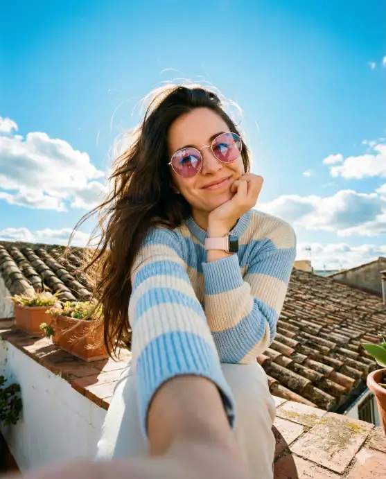 Create a bright, cheerful outdoor selfie of a young woman sitting on a rooftop ledge under a vivid blue sky filled with soft, fluffy white clouds. The camera angle is slightly low and close, as if she is holding the phone at arm's length, creating a natural wide-angle selfie perspective with subtle foreground distortion on her extended arm. She has long, dark brown hair flowing freely in the wind, strands gently lifted and illuminated by sunlight. She wears round, pink-tinted sunglasses with thin metallic frames that reflect the bright sky. Her expression is warm and genuine, with a relaxed, radiant smile. One hand supports her chin casually, giving the pose a candid, effortless feel. She is dressed in a cozy, ribbed knit sweater with horizontal pastel blue and cream stripes, paired with light-colored pants. A soft pink smartwatch or fitness band is visible on her wrist, adding a modern detail. Use aspect ratio 4:5.
