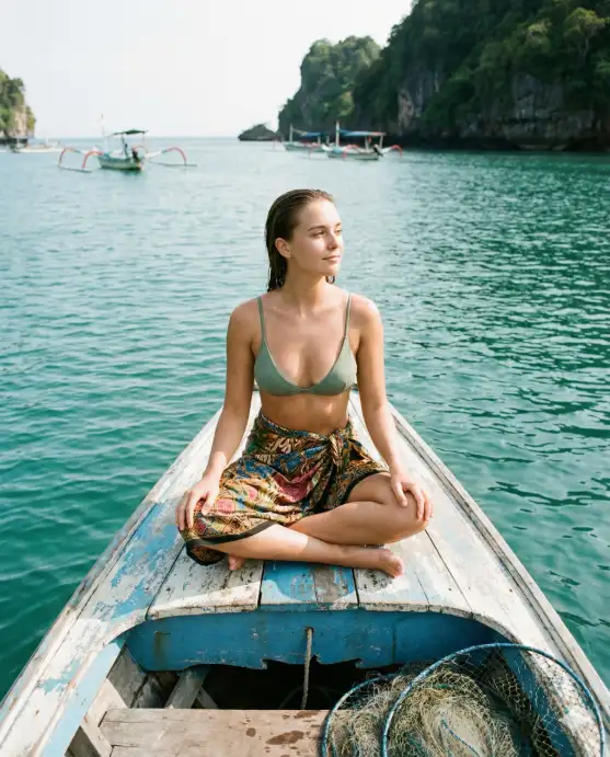 Create a serene travel photo of a young woman sitting at the front of a traditional wooden jukung boat on calm turquoise water in Bali, Indonesia. She is sitting cross-legged with her hands resting on her knees, looking out toward the horizon with a peaceful expression. She is wearing a sage green bikini top with a colorful batik sarong wrapped around her waist, and her wet hair is slicked back. The wooden boat has faded blue and white paint with visible weathering and fishing nets coiled at the back. In the distance, lush green cliffs and a few other boats dot the water. The camera is positioned at the other end of the boat, shooting toward her at a low angle. Bright natural light with turquoise and green tones. Shot with a 24mm wide-angle lens, lifestyle travel aesthetic. Use aspect ratio 4:5.