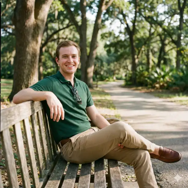 Create a photo of a man in his mid-20s with fair skin and a bright toothy smile, sitting on a park bench with one arm draped over the backrest and his body turned toward the camera in an open inviting posture, wearing a snug-fitting forest green polo shirt and classic tan chinos with brown leather loafers, clean-shaven with short dark hair neatly styled, a pair of sunglasses hooked into his collar, lush green park setting with a walking path and tall oak trees softly blurred behind him, bright natural afternoon sunlight creating warm highlights on his face with gentle dappled tree shadows, shot from eye level as a medium body shot, approachable and easygoing mood with rich greens and warm natural tones, candid outdoor portrait photography. Use aspect ratio 1:1.
