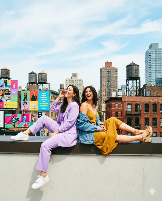 Create a photo of two female friends sitting back-to-back on a rooftop ledge with their legs stretched out, both laughing and looking away from each other. One is wearing a lavender oversized blazer with matching trousers and white sneakers, and the other is in a mustard yellow slip dress with a denim jacket draped over her shoulders and platform sandals. The background features a vibrant urban landscape with colorful billboards, water towers, and a mix of old and new architecture under a bright midday sky with wispy clouds. The camera is positioned slightly below them at a distance capturing the full scene. Bright, punchy, editorial-style color grading with vivid saturation. Shot with a 28mm lens. Use aspect ratio 4:5.