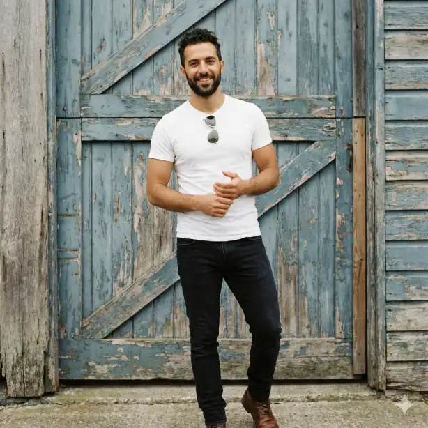 Create a portrait of a man in his early 30s with olive skin and a well-groomed dark beard, smiling softly with a relaxed open expression and eyes looking directly at the camera, standing in front of a rustic wooden barn door with peeling blue paint, hands loosely clasped in front of him in a natural comfortable stance, wearing a perfectly fitted plain white crew neck t-shirt that highlights his build and dark slim jeans with clean brown leather boots, a classic aviator sunglasses resting on top of his head, short dark hair with natural texture, soft overcast light providing even flattering illumination with no harsh shadows, shot at eye level as a medium shot from thighs up, effortlessly masculine and approachable mood with a rustic palette of white blue-grey brown and warm skin tones, natural portrait photography with a slightly desaturated vintage tone. Use aspect ratio 1:1.