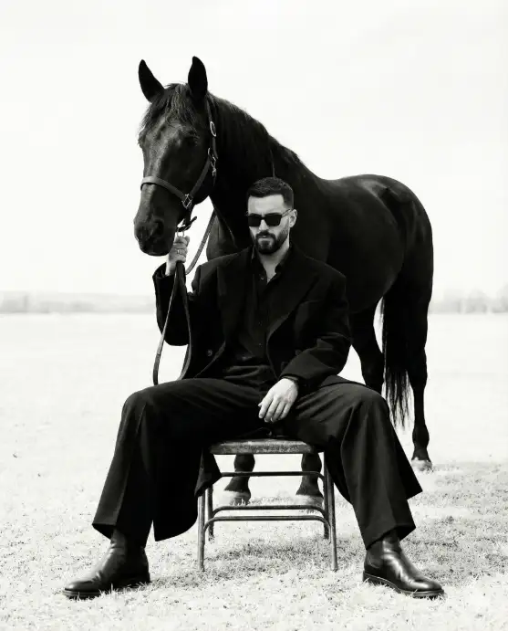 Create a striking black-and-white fashion portrait set in an open field, featuring a man seated on a simple metal chair with legs spread in a relaxed yet dominant posture. Behind him stands a large black horse, calm and imposing, positioned closely so its head hovers just over his shoulder, creating a powerful visual bond between man and animal. The horse wears a simple bridle, reins loosely held in the man's hand, symbolizing control, trust, and quiet strength. The man wears an all-black, oversized tailored coat layered over a dark shirt and wide-leg trousers, paired with polished leather boots. He has dark, styled hair, a trimmed beard, and wears dark sunglasses, concealing his eyes and adding mystery. His head tilts slightly downward, expression introspective and stoic, conveying confidence, restraint, and authority. The environment is minimal, kept intentionally understated to let the figures dominate the frame. The sky and background are bright and uncluttered, creating strong contrast with the dark silhouettes of the man and horse. Lighting is soft and natural, with even exposure and subtle highlights defining textures in fabric, skin, and the horse's coat. The image is rendered in high-contrast monochrome with a refined grain, evoking luxury editorial photography and conceptual art. The overall mood is powerful, symbolic, and cinematic. Use 4:5 aspect ratio.
