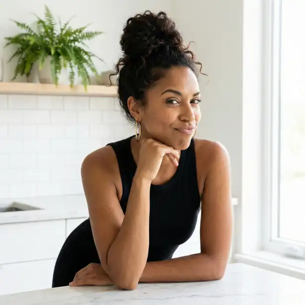 Create a photo of a woman in her early 30s with warm brown skin and defined jawline, giving a playful smirk with one eyebrow slightly raised, leaning forward with her elbows resting on a marble countertop and her chin propped on one hand, wearing a fitted black ribbed tank top and oversized gold hoop earrings, her dark curly hair pulled up in a messy high bun with a few loose tendrils framing her face, bold red lipstick as the only makeup statement, set in a modern minimalist kitchen with white subway tiles and a green plant in the background, bright soft natural light streaming from a large window to the right, shot from a slightly elevated angle as a medium close-up, flirty and self-assured mood with a clean palette of black white and pops of gold and green, candid editorial photography. Use aspect ratio 1:1.
