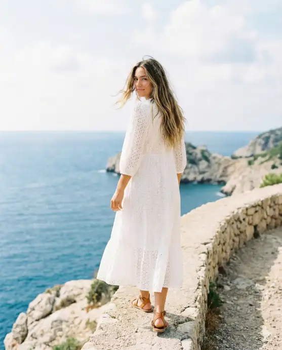 Create a photo of a young woman walking along a narrow stone path on a coastal cliff, looking back over her shoulder toward the camera with a relaxed smile. She is wearing a flowy white broderie anglaise midi dress with thin straps and a slightly open neckline, and tan leather flat sandals. Her hair is long and loose, slightly windswept. The background shows a dramatic blue sea, rocky coastline, and a pale blue sky with soft white clouds. The camera is at a low angle slightly behind her, capturing her in motion. Bright, clean natural light with airy, sun-bleached tones. Fashion lifestyle photography. Use aspect ratio 4:5.