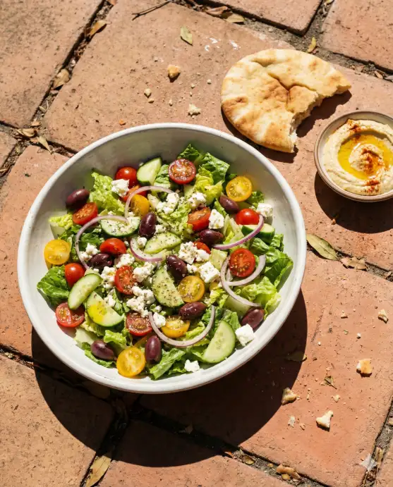 Create a realistic photo of a large white ceramic bowl filled with a Mediterranean salad on a terracotta tile surface outdoors. The salad contains chopped romaine, cherry tomatoes halved to show the juicy interior, sliced cucumber, Kalamata olives, crumbled feta cheese, thin red onion rings, and a generous drizzle of olive oil with dried oregano visible. A piece of warm pita bread torn in half sits beside the bowl, along with a small dish of hummus with a swirl of olive oil and paprika on top. The camera is at a 45-degree angle from slightly above. Bright, direct midday sunlight casting hard, defined shadows on the surface, giving it a raw Mediterranean summer feel. Warm, sun-bleached color palette with pops of red and green. Shot with a 35mm lens. Use aspect ratio 4:5.