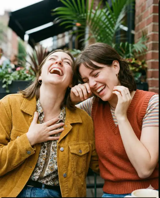 Create a candid laughing portrait of two friends captured mid-laughter, standing close together with casual body language and expressive gestures. Their posture is dynamic, slightly leaning toward each other. They are wearing trendy casual outfits with complementary colors. Shot at eye level with shallow depth of field and natural daylight. Background is an outdoor lifestyle setting with soft blur and warm tones, giving a spontaneous Instagram photo feel. Use aspect ratio 4:5.