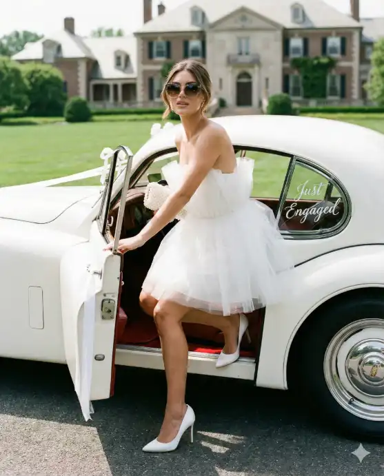 Create a photo of a bride-to-be stepping out of a vintage white car, one heel touching the ground, one hand on the car door, wearing a white strapless cocktail dress with a tulle overlay, oversized sunglasses, a pearl clutch in her other hand, confident glamorous expression, the car decorated with "Just Engaged" written on the rear window and white ribbons, green lawn and a grand estate house in the background, bright daylight, shot from a low angle to the side, full body composition, 50mm lens, fashion editorial photography style. Use aspect ratio 4:5.