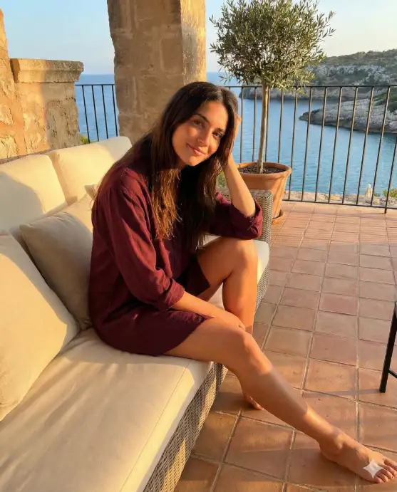 Create a cinematic golden-hour portrait of a young woman seated on a sunlit terrace overlooking a serene coastal landscape. She sits on a warm-toned tiled floor, leaning gently against a cream-colored outdoor sofa with soft cushions. Her posture is relaxed and intimate, one knee bent toward her chest, the other leg extended naturally, one arm resting loosely over her knee while the other supports her head. She wears a deep burgundy or wine-colored long-sleeve shirt dress, slightly oversized, with soft fabric that catches the warm light. The dress drapes naturally over her frame, enhancing the relaxed, effortless aesthetic. Her long, dark brown hair falls freely over her shoulders, illuminated by the low golden sun, creating subtle highlights and depth. The sunlight streams in from the side, casting strong, warm shadows and dramatic light patterns across her face, legs, and the surrounding architecture. The lighting should feel rich and cinematic, evoking late afternoon or early sunset. Behind her, a black metal balcony railing frames a distant view of calm blue ocean waters and a rugged coastline. A potted olive tree stands nearby, its leaves casting soft, organic shadows on the warm-toned walls. The overall setting suggests a Mediterranean-inspired terrace with earthy textures and natural materials. Use aspect ratio 4:5.