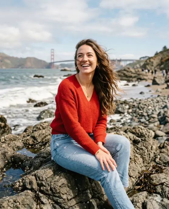 Create a vibrant outdoor lifestyle portrait of a young woman sitting casually on rugged seaside rocks near a shoreline, smiling joyfully while looking slightly upward as a gentle coastal breeze flows through her long hair, strands moving naturally across her face to create a carefree candid moment, wearing a bright red V-neck sweater paired with light blue denim jeans and small gold hoop earrings, her posture relaxed with shoulders slightly forward as she leans comfortably, the background featuring a rocky beachscape and a distant bridge softly blurred by shallow depth of field, captured in soft natural daylight with warm tones and subtle wind movement that gives the image a cinematic, carefree coastal mood. Use aspect ratio 4:5.