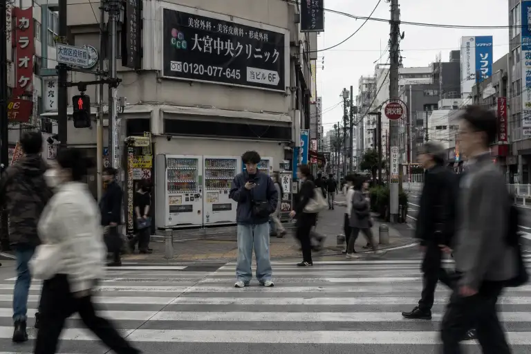 Create a street photo of a young man standing at a busy intersection in Tokyo, waiting for the light to change. He is wearing a navy blue windbreaker jacket, baggy light-wash jeans, and retro running sneakers with a small crossbody bag slung across his chest. He is looking down at his phone, unaware of the camera. Pedestrians are blurred in motion around him while he stands still in the center of the frame. The background shows tall buildings covered in Japanese signage, vending machines, and power lines against an overcast sky. The camera is across the street at chest height, capturing the full scene. Slightly desaturated tones with muted blues and grays. Shot with a 28mm lens, documentary street photography style. Use aspect ratio 3:2.