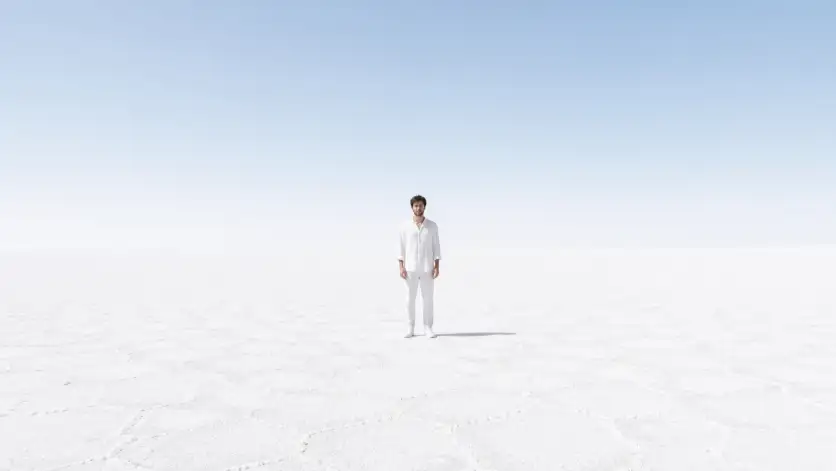 Create a minimalist photo of a man standing alone in the center of a vast white salt flat that stretches to the horizon in every direction. He is wearing an all-white outfit consisting of a loose white linen shirt, white linen trousers, and white canvas sneakers. He is standing still with his arms at his sides, facing the camera with a calm, neutral expression. The sky above is a smooth gradient from pale blue at the top to nearly white at the horizon, blending seamlessly with the white ground. The camera is at a distance, capturing his full body as a small figure in the expansive white landscape. Ultra-clean, high-key lighting with soft shadows. Minimal color palette of white, light gray, and pale blue. Shot with a 24mm lens. Use aspect ratio 16:9.