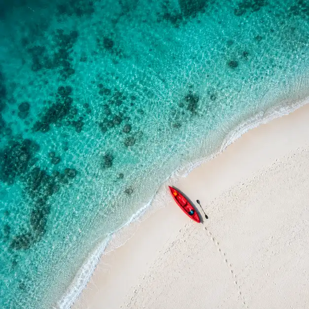 Create an aerial photo shot directly from above looking straight down at a turquoise ocean meeting a white sandy beach. A single red kayak is pulled halfway onto the sand with a paddle resting beside it. Footprints lead from the kayak across the sand and disappear into the water. The ocean water is crystal clear near the shore, showing the sandy bottom and a few dark patches of coral beneath the surface, transitioning to deeper turquoise further out. Small white waves are breaking gently along the shoreline. The composition is geometric with the diagonal line of the beach cutting across the frame. Bright, vivid tropical colors with strong contrast between the red kayak, white sand, and turquoise water. Shot with a wide-angle lens from a drone at 50 meters altitude. Use aspect ratio 1:1.