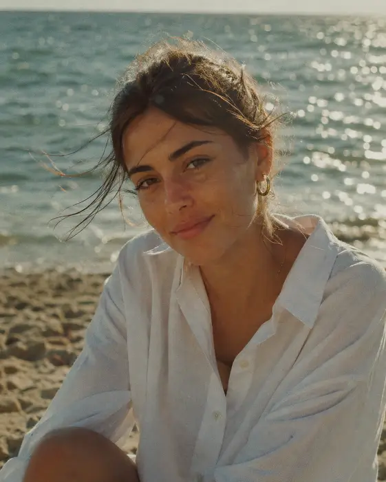 Create a film-style portrait of a woman in her mid 20s sitting on a beach at late afternoon, looking softly into the camera with a relaxed half-smile. She is wearing a slightly oversized crisp white button-up shirt with the top two buttons undone and small gold hoop earrings. Her dark hair is pulled back loosely with a few strands blowing across her face from the sea breeze. Shoot at eye level with the ocean filling the entire background, sunlight glittering on the water in soft sparkles. Use warm afternoon light with visible film grain, slight color shift toward green and amber, and a soft analog haze. The mood should feel quiet, candid, and effortlessly Instagram. Use aspect ratio 4:5.