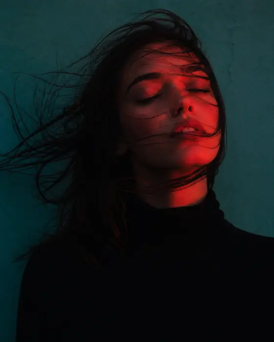 Create a moody close-up portrait of a young woman with her eyes closed and chin slightly lifted, standing against a flat teal-painted wall. She is wearing a simple black turtleneck and no visible jewelry. Her dark hair is being blown across her face by a strong side wind, with strands wrapping around her cheek and neck. Light a deep red gel light onto her face from one side so half her skin glows red while the rest stays in shadow. Shoot at eye level, tightly framed from the chest up. Add fine grain and a slightly grainy analog texture. The mood should feel dreamy, editorial, and emotional. Use aspect ratio 4:5.