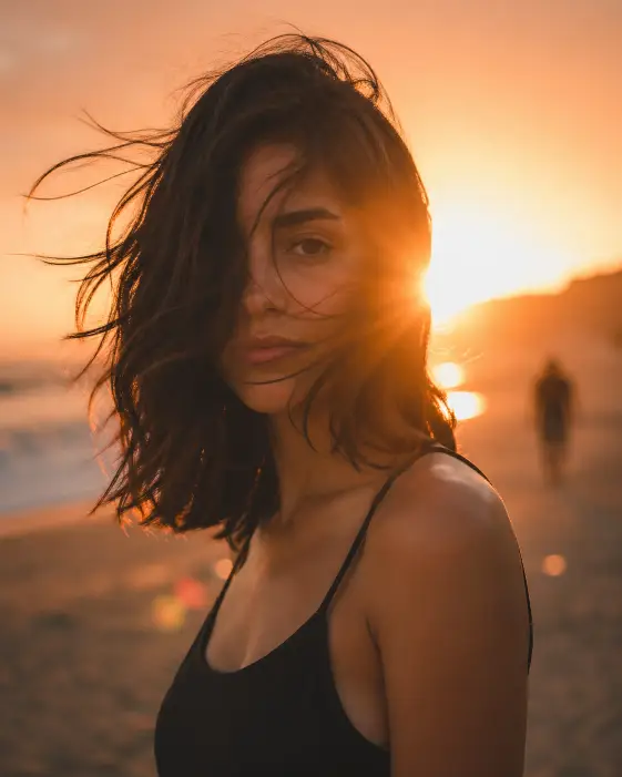 Create a backlit portrait of a young woman standing on a beach at sunset, with the sun setting directly behind her right shoulder and creating a strong lens flare. She is wearing a simple black tank top with thin straps and no jewelry. Her shoulder-length dark hair is windswept, with several strands falling across half of her face so only one eye is fully visible. She looks calmly toward the camera with a soft, neutral expression. Shoot at eye level with a 50mm lens, shallow depth of field. In the soft blurred background include a faint silhouette of another person walking far behind her and warm bokeh from the setting sun. Use warm orange and amber tones with cinematic color grading. Use aspect ratio 4:5.