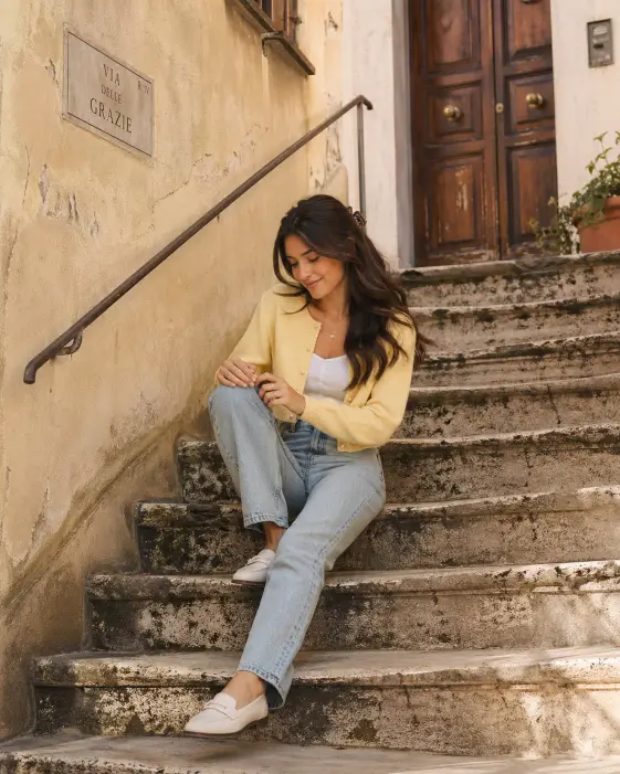 Create a soft street-style portrait of a young woman sitting on a worn stone staircase outside an old European apartment building, with one knee pulled up and the other leg stretched down a few steps. She is wearing a soft butter-yellow cropped cardigan with small pearl buttons over a plain white camisole, paired with high-waisted light wash jeans and white leather loafers. Her dark hair is in loose waves falling past her shoulders with a small claw clip holding back one side. She is looking down at her hands resting on her knee with a soft natural smile. Shoot from a low angle at the bottom of the stairs to capture the full staircase leading up to her. The background shows a weathered cream stucco wall and a small wooden door slightly out of focus. Use warm afternoon light with soft shadows from the staircase, gentle film grain, and a vintage European street-style mood. Use aspect ratio 4:5.