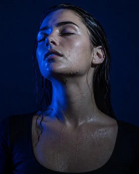 Create a moody editorial close-up portrait of a young woman with her eyes closed and her face tilted slightly upward, water droplets clinging to her skin and her dark hair slicked back wet. She is wearing a simple plain black scoop neck top. Light a deep cobalt blue gel light onto her face from one side and a soft cool white fill from the other, leaving deep shadows in the contours. Shoot tightly cropped from the chest up against a flat dark navy background. Use cold blue tones with rich saturation, fine analog grain, and a calm, surrendered, almost-underwater feeling. Use aspect ratio 4:5.