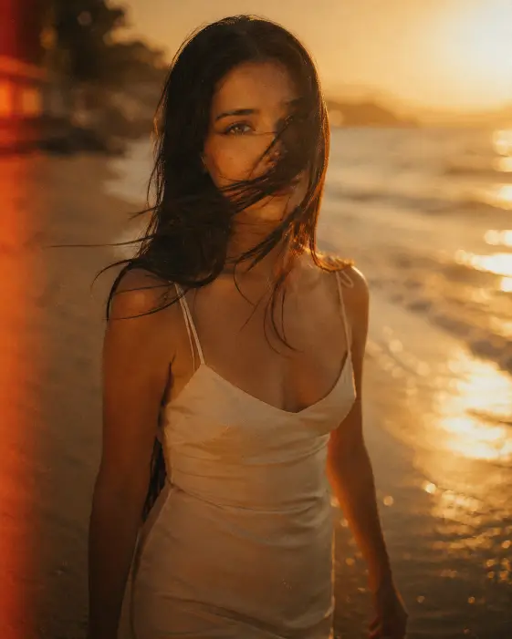 Create a film-style portrait of a young woman walking slowly along the edge of a beach at sunset, captured in the moment a strong gust of wind wraps a long strand of her dark hair across her face. She is wearing a soft cream slip dress with thin straps. Her arms are relaxed at her sides and she is looking softly toward the camera with one eye half-covered by her hair. Shoot at eye level with a shallow depth of field. The sun is low and just out of frame on the right, creating a strong warm side glow on her cheek and a soft golden flare. The background is a hazy out-of-focus ocean with warm sparkles on the water. Use warm honey and amber tones with heavy analog film grain and slight light leaks. Use aspect ratio 4:5.