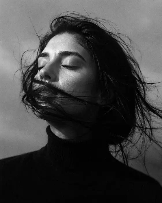 Create a black and white film-style close-up portrait of a young woman with her eyes closed and her chin slightly raised, strong wind blowing her dark hair across the lower half of her face. She is wearing a simple black turtleneck. Shoot tightly cropped from the shoulders up at eye level against a plain hazy gray sky background. Use dramatic side lighting from a soft natural source, creating bright highlights on her cheekbone and deep shadows in the hollows. Use rich black and white tonality with deep blacks, soft midtones, and visible 35mm analog grain. The mood should feel quiet, emotional, and editorial. Use aspect ratio 4:5.