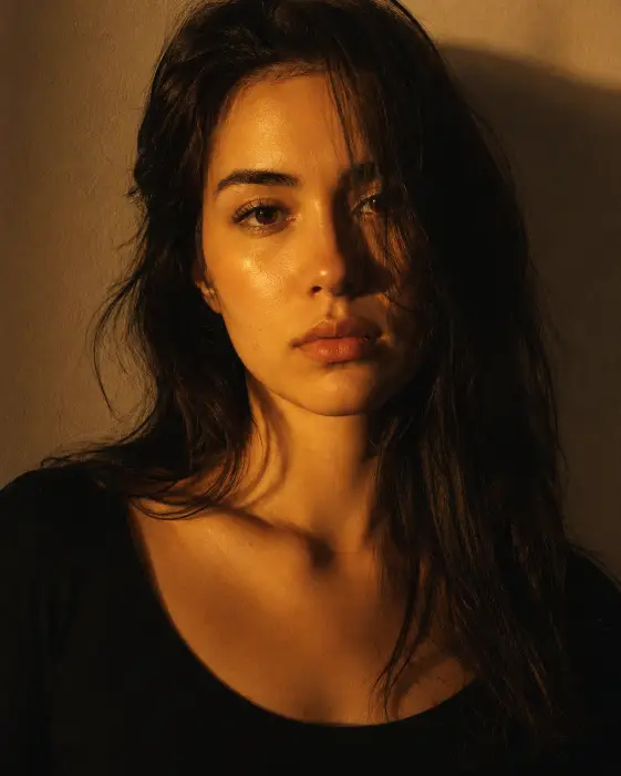 Create a moody editorial close-up portrait of a young woman standing against a plain warm beige wall, looking directly into the camera with a calm, neutral expression. She is wearing a fitted plain black scoop neck top and no jewelry. Her dark hair is loose with a few strands falling across one side of her face. Light a soft warm yellow gel light onto her face from one side so half of her skin glows golden yellow while the other side falls into deep warm shadow. Shoot tightly cropped from the chest up at eye level. Use rich warm tones, fine analog film grain, and a quiet, magazine-cover editorial mood. Use aspect ratio 4:5.