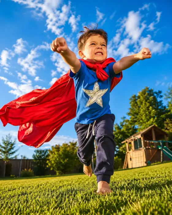 Create a fun, energetic outdoor photo of a 5-year-old boy running across a green backyard lawn with his arms stretched forward like he is flying. He is wearing a homemade superhero cape made from a bright red bedsheet tied around his neck over a blue t-shirt with a hand-drawn star on the chest, paired with dark sweatpants and bare feet on the grass. His face shows fierce determination with squinted eyes and a clenched jaw. His cape is billowing behind him in the wind. The camera is at a low angle, close to the ground, shooting upward to make him look larger than life against a bright blue sky with fluffy white clouds. Vibrant, saturated colors with warm afternoon sunlight casting long shadows on the grass. Shot with a 24mm wide-angle lens, fast shutter speed to freeze motion, editorial lifestyle aesthetic. Use aspect ratio 4:5.