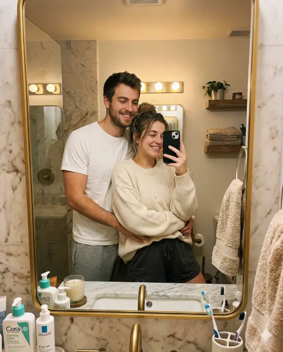 Create a realistic couple mirror selfie of a young man and woman standing in front of a large bathroom mirror in the morning. The man is standing behind the woman with one arm wrapped around her waist, holding the phone with his other hand to take the selfie. He is wearing a plain white t-shirt and gray sweatpants, and she is wearing an oversized cream knit sweater and shorts with her hair in a messy bun. Both are smiling naturally with sleepy, relaxed expressions. The bathroom has warm vanity lighting, a marble countertop with skincare bottles, and a soft beige towel hanging on the side. The mirror has a thin gold frame. The camera is at chest level, shot through the mirror reflection, with soft warm tones and a cozy morning atmosphere. Use aspect ratio 4:5.