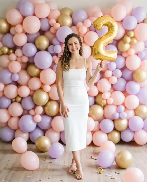 Create a photo of a young woman standing in front of a large wall of pastel-colored balloons in shades of blush pink, lavender, and soft gold. She is wearing a fitted white midi dress with thin straps and delicate gold hoop earrings. Her hair is styled in loose curls falling over one shoulder. She is holding a single gold foil number balloon in one hand and smiling warmly at the camera with her other hand resting lightly on her hip. The camera is at eye level, shooting straight on. Soft, diffused indoor lighting with no harsh shadows. The floor is light wood, and a few balloons have fallen to the ground around her feet. Warm, cheerful, lifestyle portrait style. Shot with an 85mm lens, shallow depth of field. Use aspect ratio 4:5.
