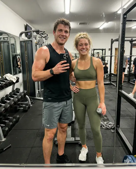 Create a realistic couple mirror selfie of a young athletic man and woman standing in front of a full-length gym mirror after a workout. The man is wearing a fitted black tank top and gray athletic shorts, flexing one arm slightly while holding the phone with the other. The woman is standing beside him in a matching olive green sports bra and high-waisted leggings with white training shoes, one hand on her hip and the other holding a water bottle. Both have light sweat on their skin and natural, proud expressions. The background shows gym equipment including dumbbells, a cable machine, and rubber flooring. Harsh overhead fluorescent lighting creates sharp, clean shadows. The mood is energetic and authentic. Shot at waist level through the mirror. Use aspect ratio 4:5.