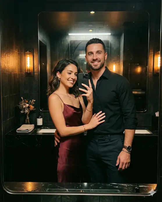 Create a realistic couple mirror selfie of a man and woman in a dimly lit upscale restaurant restroom. The man is wearing a fitted black button-down shirt with rolled sleeves and dark trousers, standing slightly behind the woman with his hand on her lower back. The woman is wearing a burgundy satin slip dress with thin straps and small gold hoop earrings, holding the phone up with one hand while the other rests on his chest. Both are looking into the mirror with confident, relaxed smiles. The restroom has dark tiled walls, a large frameless mirror, warm amber wall sconces, and a sleek black countertop with a small vase of dried flowers. The image has a moody, warm color palette with rich shadows. Shot at chest level through the mirror reflection. Use aspect ratio 4:5.