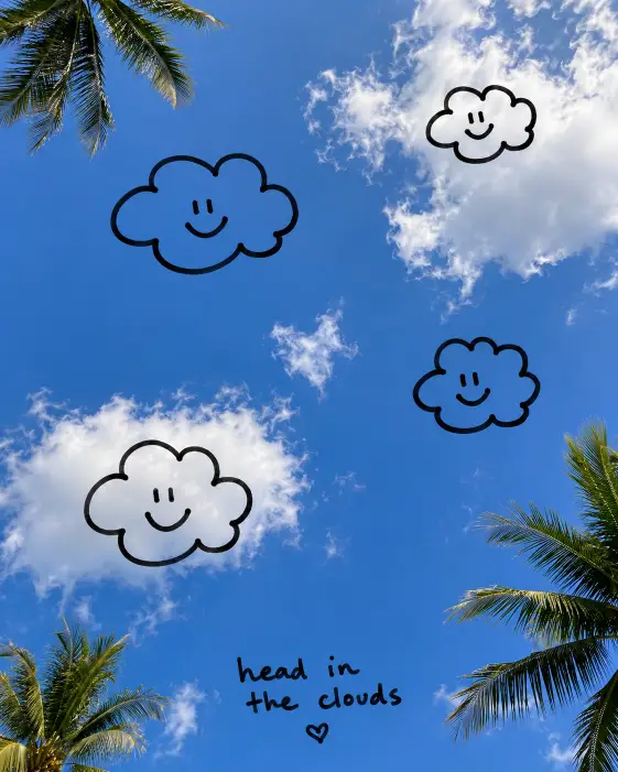 Create a vertical photo looking straight up at a clear blue sky with a few fluffy white clouds drifting across it. The camera angle is directly overhead, as if lying flat on the ground and pointing the camera up. Lighting is bright midday sun. The composition is mostly sky, with the edge of a few palm tree leaves peeking in from the corners. Add hand-drawn doodle clouds with simple smiley faces inside them, plus a small scribbled caption "head in the clouds" near the bottom of the frame in handwritten script. The doodles should look like they were drawn with a chunky black marker over the printed photo. Use aspect ratio 4:5.