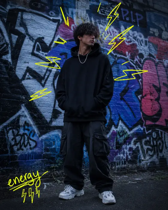 Create a vertical photo of a young man standing in front of a graffiti-covered brick wall, hands in his hoodie pockets and looking off to the side with a serious expression. He's wearing an oversized black hoodie, baggy cargo pants, white chunky sneakers, and a silver chain necklace. His hair is messy and slightly covers his forehead. The camera angle is at chest height, straight on. Lighting is moody with cool blue and gray tones. The background is a colorful graffiti wall with bold tags and shapes. Add hand-drawn neon yellow lightning bolt doodles around his shoulders and head, plus a small scribbled caption "energy" near the corner in jagged handwriting. The doodles should look like marker drawings on top of a printed photo. Use aspect ratio 4:5.