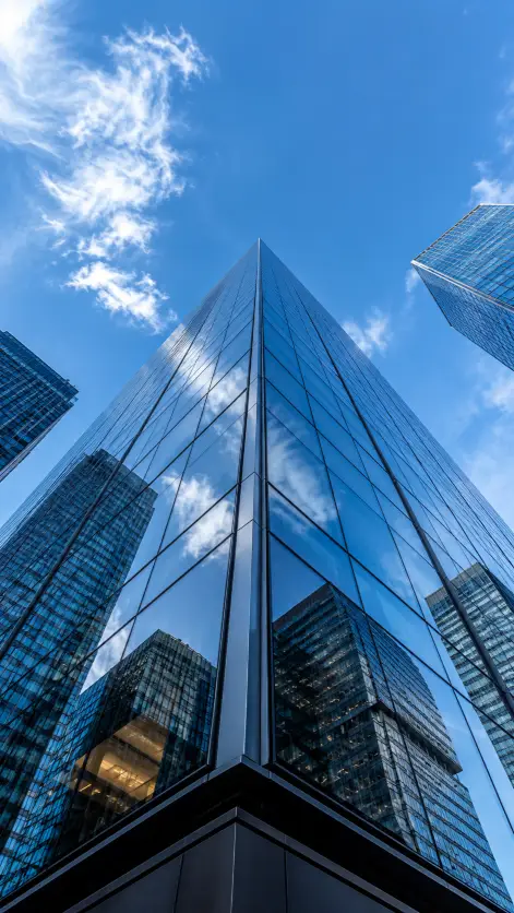 Create a worm's eye view photo looking straight up at a tall glass skyscraper in a downtown business district. The camera is positioned at the very base of the building pointing skyward, so the building's edges converge dramatically toward the center of the frame. Reflections of clouds and other buildings shimmer across the glass facade. A clear blue sky with a few wispy white clouds fills the top of the frame. Sharp, crisp architectural photography style with high contrast and rich blues. Use aspect ratio 9:16.