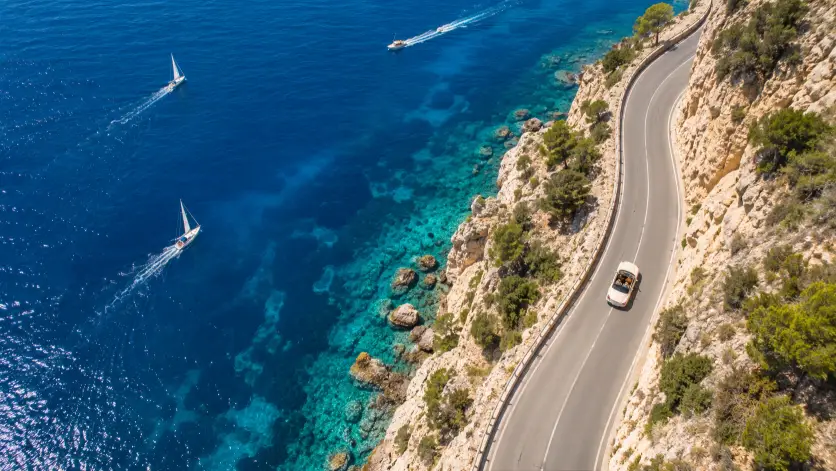 Create a bird's eye aerial photo of a winding coastal road cutting through cliffs along the Mediterranean coast. A single vintage cream-colored convertible is driving along the road, with crystal turquoise water on one side and rocky beige cliffs on the other. Small sailboats dot the water in the distance, leaving thin white wakes behind them. Shoot from directly above at a high altitude as if from a drone. The lighting is bright midday sun with rich saturated blues and warm sandy tones. Use aspect ratio 16:9.