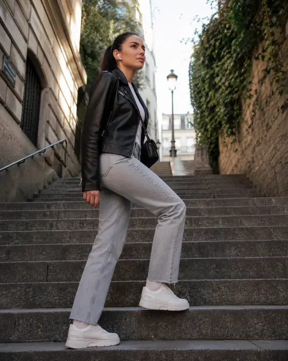 Create a photo of a young woman climbing a wide outdoor stone staircase in the city, captured mid-step with her right foot on the upper stair and the other still descending below. She is wearing a black leather jacket, white t-shirt, light gray straight-leg jeans, and white chunky sneakers, with white wireless earbuds in her ears and a small black crossbody bag. Her hair is in a sleek high ponytail and her expression is focused and confident, eyes locked ahead. The staircase is flanked by old stone buildings and ivy-covered walls, with soft morning light spilling down from above. The camera is at a low angle on the stairs below her, shooting upward to emphasize the climb. Crisp natural light with cool urban tones. Shot with a 35mm lens, modern lifestyle style. Use aspect ratio 4:5.