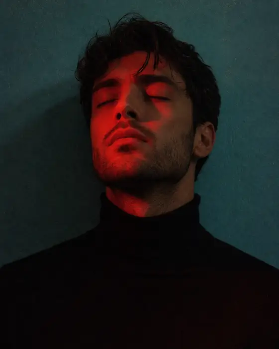 Create a moody close-up portrait of a young man with his eyes closed and chin slightly lifted, standing against a flat teal-painted wall. He is wearing a simple black turtleneck and no jewelry. His dark hair is short and slightly tousled, with a few strands falling on his forehead. He has a clean stubble beard along his jaw. Light a deep red gel light onto his face from one side so half his skin glows red while the rest stays in shadow. Shoot at eye level, tightly framed from the chest up. Add fine grain and a slightly grainy analog texture. The mood should feel dreamy, editorial, and emotional. Use aspect ratio 4:5.