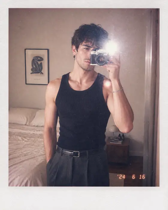 Create a polaroid-style mirror selfie of a young man standing in a softly lit minimalist bedroom, holding a vintage 35mm film camera up to his face so it partially covers his right eye. He is wearing a fitted plain black ribbed tank top tucked into high-rise dark gray trousers with a thin black leather belt, and a silver chain bracelet on his right wrist. His short dark hair is slightly messy with a natural texture and he has a clean light stubble. The camera flash creates a bright soft white reflection in the mirror and a glow on his shoulder. The bedroom behind him shows a neatly made bed with cream linen, a small wooden nightstand, and a single framed art print on a beige wall. Add visible film grain, slight overexposure, a faded polaroid color tone with a subtle pink and cream cast, and a soft date stamp in the bottom right corner. Use aspect ratio 4:5.