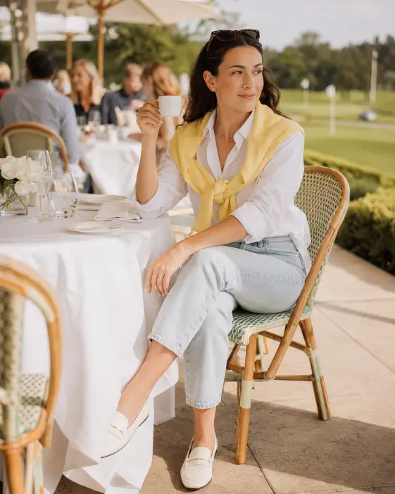 Create a photo of a woman sitting at a round outdoor table at a country club brunch, one hand holding a small espresso cup and the other resting on the white linen tablecloth. She is wearing a pale yellow cotton sweater knotted over her shoulders on top of a white button-down shirt, light wash cropped jeans, and white leather loafers. Small pearl studs and a delicate gold chain complete the look. The camera is at eye level, capturing her from a slight three-quarter angle. The background shows other diners blurred softly, white umbrellas, a manicured lawn, and golf flags in the far distance. Bright natural midday light with warm cream tones. Shot with a 50mm lens, shallow depth of field. Use aspect ratio 4:5.