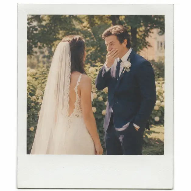 Create a Polaroid-style wedding photo of a bride and groom during their first look, the groom turning around to see the bride with his hand over his mouth in surprise. The bride is wearing a fitted ivory lace gown with a long veil, and the groom is in a classic navy suit with a white boutonniere. They are standing close together in a sunlit garden with soft greenery behind them. Shoot at eye level with a slightly soft focus. Warm afternoon light gives the image a gentle glow, and the photo has a faded vintage tint with visible film grain and a thick white Polaroid border. Use aspect ratio 1:1.