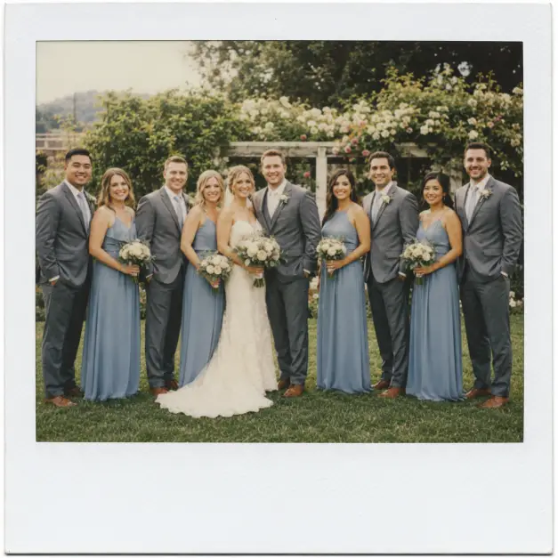 Create a Polaroid-style wedding photo of a bride and groom standing in the center of their bridal party, with bridesmaids in matching dusty blue dresses and groomsmen in grey suits gathered around them, all smiling. They are standing on green grass with a soft blurred garden venue behind them. The camera is at eye level in a wide shot capturing the full group. Bright natural daylight with a warm vintage tone, soft film grain, and a thick white Polaroid border. Use aspect ratio 1:1.