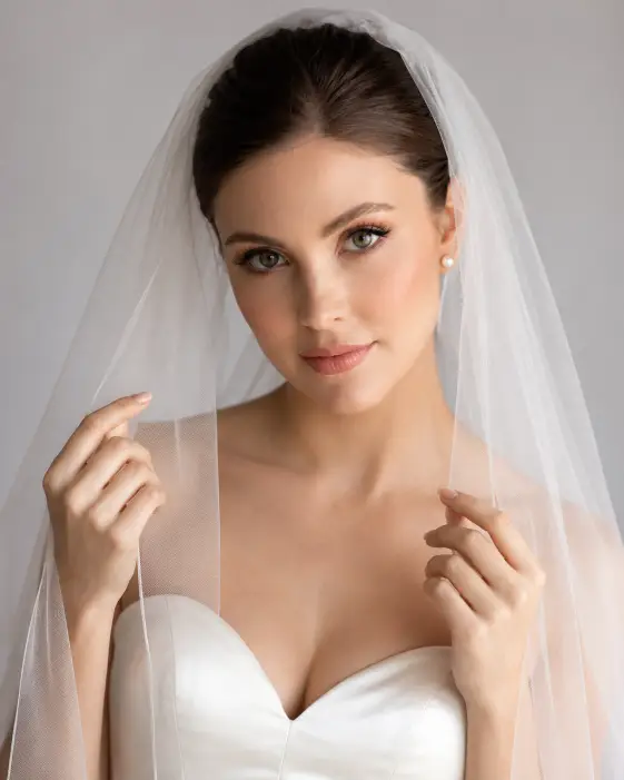Create an elegant bridal portrait of a young woman wearing a long sheer veil draped over her head and shoulders. She holds the edges of the veil gently with both hands near her face, looking directly into the camera with a soft, serene expression. She wears a fitted satin wedding gown with a sweetheart neckline. Her makeup is refined with soft rosy tones and her hair is pulled back neatly beneath the veil. The camera is at eye level in a close-up framing, focused on her face and the delicate fabric of the veil. Soft, even studio lighting with a gentle glow. The background is a clean, softly blurred light gray. Shot with an 85mm portrait lens, creamy bokeh. Use aspect ratio 4:5.